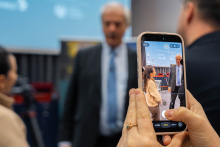 Osoba trzyma smartfon i fotografuje uczestnika wydarzenia. Na ekranie telefonu widać fotografowaną osobę oraz planszę „The Nobel Prize. Nobel Prize Inspiration Initiative”.
