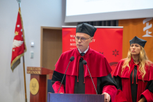 Prodziekan w stroju akademickim przemawia przy mównicy podczas inauguracji roku akademickiego. Za nim stoi prodziekanka. W tle znajduje się ekran prezentacyjny, sztandar uczelni oraz ścianka w kolorze bordowym.
