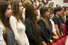 Grupa studentów stoi między rzędami siedzeń w auli i uczestniczy w uroczystości.