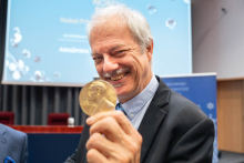 Uczestnik wydarzenia trzyma przed sobą medal i uśmiecha się do aparatu. W tle znajduje się ekran z napisem „Nobel Prize Inspiration Initiative” oraz baner Warszawskiego Uniwersytetu Medycznego.