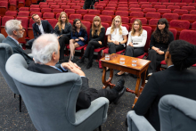 Dziewięć osób uczestniczy w rozmowie w auli. Siedem osób siedzi w półokręgu naprzeciw dwóch osób w fotelach. Między uczestnikami stoi stolik ze szklankami wody.
