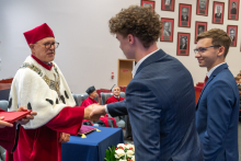 Dwóch młodych mężczyzn odbiera od władz dokumenty. Jeden mężczyzna podaje rękę przedstawicielowi władz uczelni ubranemu w togę.