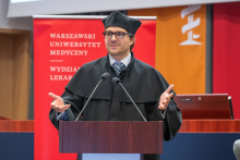 Młody mężczyzna ubrany w czarną togę akademicką stoi za mównicą i przemawia. W tle widać bordową ściankę z logo wydziału oraz fragment dekoracji.