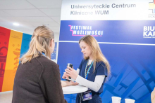 Pracownica w granatowej bluzce przy stanowisku Uniwersyteckiego Centrum Klinicznego WUM skanuje lub obsługuje urządzenie medyczne podczas rozmowy z uczestniczką targów.