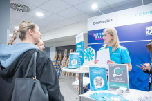 Uśmiechnięta przedstawicielarka firmy Cosmedica w niebieskiej koszulce rozmawia z uczestniczkami targów przy stoisku informacyjnym z materiałami rekrutacyjnymi.