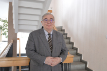 Person wearing glasses and a grey suit, standing with hands clasped beside an indoor staircase.