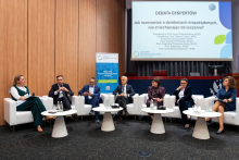 Seven participants in an expert debate during the Pharmacovigilance Polska 2026 Congress. The participants are seated in white armchairs in front of a screen displaying the debate topic on adverse reactions and treatment.