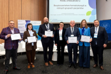 Seven people posing for a group photo after a panel discussion on the safety of pharmacotherapy in different patient groups. Five of them are holding framed certificates.