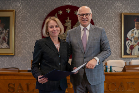 Kobieta i mężczyzna stojący obok siebie. Kobieta po lewej ubrana jest w ciemny garnitur i trzyma przed sobą niebieską teczkę. Mężczyzna po prawej ma na sobie szary garnitur w kratę oraz jasnofioletowy krawat. Uśmiechają się do zdjęcia. Za nimi widać drewniany stół. W tle znajduje się czerwone godło WUM z orłem i gwiazdami. 
