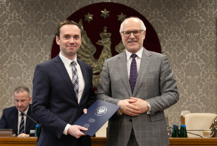 W przestronnej sali stoją obok siebie dwaj mężczyźni. Uśmiechają się do zdjęcia. Mężczyzna po lewej ma ciemny garnitur i krawat i trzyma niebieską teczkę z godłem i nazwą WUM. Mężczyzna po lewej ma szary garnitur i ciemny krawat. W tle widoczny jest duży herb z orłem i gwiazdami na czerwonym tle oraz osoby siedzące przy stole. 