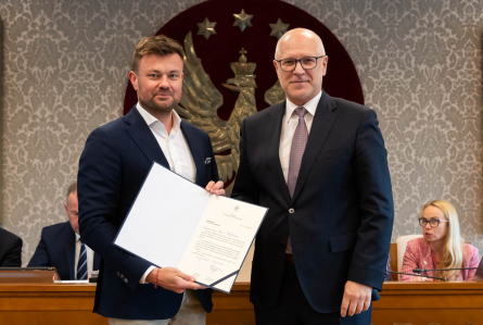 Dwóch mężczyzn w ciemnych garniturach stoi obok siebie w sali konferencyjnej. Mężczyzna po prawej ma okulary i krawat. Mężczyźni uśmiechają się do zdjęcia. Jeden z nich trzyma otwartą teczkę z podpisanym dokumentem. W tle widoczny jest herb z orłem oraz osoby siedzące przy stole.