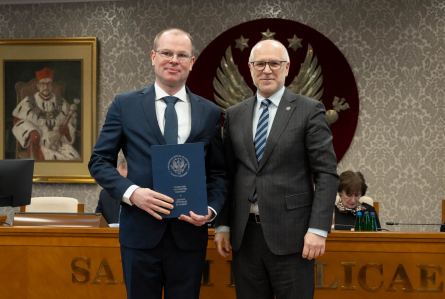 Dwóch mężczyzn stoi obok siebie, są elegancko ubrani. Jeden z nich trzyma teczkę z otwartym dokumentem. W tle siedzą dwie osoby oraz widać duże godło z orłem. 