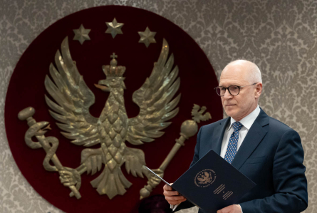 Mężczyzna stoi przy mównicy i odczytuje dokument. W tle widoczny jest herb uczelni - złoty orzeł na czerwonym tle. 