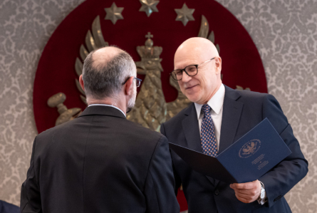 Dwóch mężczyzn stoi naprzeciw siebie w sali senatu WUM. Jeden wręcza drugiemu otwartą teczkę z dokumentem. W tle widoczny jest herb uczelni.