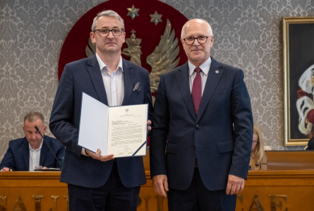 Dwie osoby stoją przed stołem prezydialnym. Jedna z nich trzyma otwartą teczkę z dokumentem skierowanym w stronę aparatu. W tle widoczne są osoby uczestniczące w posiedzeniu oraz godło na ścianie.