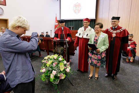 Trzy osoby - dwóch mężczyzn i kobieta stoją na środku auli. Przed nimi stoi mężczyzna, który wykonuje fotografię. 