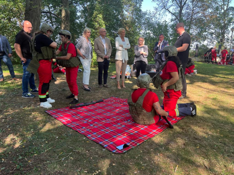 Na polanie rozłożony koc. Na nim siedzi kobieta w kasku i kamizelce kuloodpornej. Obok niej klęczy druga w takim samym stroju. Wokół stoi kilka osób, które się temu przyglądają. Wśród nich wysoka kobieta, blondynka w białej marynarce i beżowych spodniach.