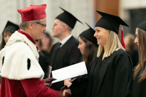 Grupa osób ubranych w stroje akademickie. Mężczyzna ubrany w czerwono-białą togę rektorską podaje dyplom uśmiechniętej kobiecie, ubranej w czarną togę i biret.