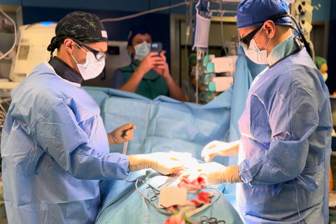 Dwóch lekarzy, ubranych w stroje chirurgiczne, w czepkach i maseczkach stoi nad pacjentem i przeprowadza zabieg.