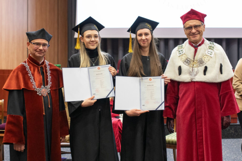 Na zdjęciu cztery osoby, po lewej i po prawej dwóch mężczyzn, w środku - dwie młody kobiety. Kobiety są ubrane w czarne togi i birety, trzymają w rękach dyplomy.