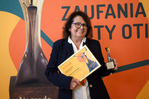 Na zdjęciu prof. Lidia Rudnicka, kobieta w średnim wieku, w okularach, elegancko ubrana. W rękach trzymaa dyplom i statuetkę. Dyplom jest żółty, statuetka jest srebrna z czarną podstawą, na której znajduje się tabliczka z napisem "Nagroda Zaufania Złoty OTIS". W tle widoczna jest pomarańczowo-żółtka ścianka z fragment dużej statuetki.