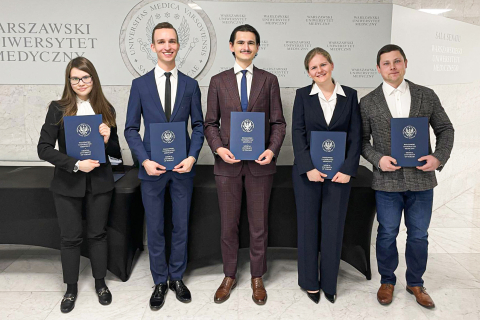 Studenci i absolwenci z nagrodami rektora za działalność naukową. Grupa młodych osób, elegancko ubranych, pozują do zdjęcia z granatowymi teczkami w dłoniach.