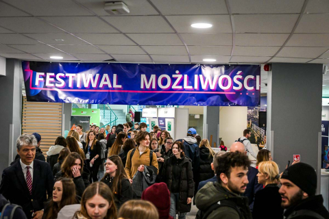 Tłum ludzi, uczestników wydarzenia Festiwalu Możliwości. Znajdują się na korytarzu, na którym są też różne stoiska wystawiennicze. Nad nimi wisi fioletowy baner z napisem Festiwal Możliwości. 