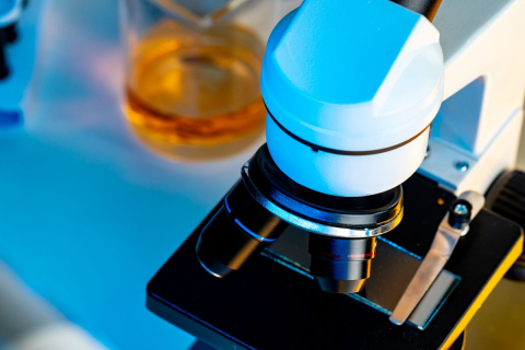 Mikroskop laboratoryjny z trzema obiektywami, ustawiony na stole. W tle znajduje się szklane naczynie z żółtą cieczą. Całość jest oświetlona niebieskim i żółtym światłem, co nadaje zdjęciu kontrastowy wygląd.