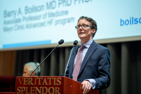 A middle-aged man, wearing glasses. He is wearing a dark suit, a light blue shirt and a light pink tie. He stands behind a lectern and speaks. The rostrum is brown and has the inscription “VERITATIS SPLENDOR” in gold. In the background is a multimedia screen with text.