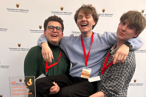 Three young men - students of WUM against the background of a wall with the inscription “Medical University of Silesian Piasts in Wroclaw”. The man in the center is held up by the men standing on the right and left. They are all smiling. Around their necks they have red lanyards with badges. The person on the left is wearing a green sweater, the person in the middle is wearing a blue sweater, and the person on the right is wearing a grayish sweater. In the foreground is an award with the words “Diagnostic S