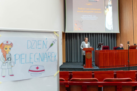 Wnętrze auli z drewnianymi panelami na ścianach i rzędami czerwonych foteli. Na pierwszym planie, po lewej stronie, widoczny jest plakat z napisem „DZIEŃ PIELĘGNIAREK”. Plakat zawiera rysunek pielęgniarki w białym uniformie, ze stetoskopem na szyi i pielęgniarską czapką z czerwonym krzyżem. Obok napisu znajduje się ilustracja strzykawki. W głębi zdjęcia, na scenie, stoi kobieta przemawiająca zza mównicy. Ma jasną marynarkę, okulary i korale.