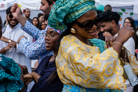 A group of people dancing during a festival. In the center, a woman in a yellow dress with a pattern and a turquoise headdress, with jewelry. She has dark glasses over her eyes and gold earrings in her ears. In the background are colorfully dressed people, tents and festival decorations.