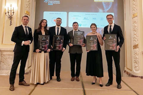 Sześć elegancko ubranych osób. Wszyscy uśmiechają się do zdjęcia. Pięć z nich trzyma w rękach dyplomy. Stoją w jasnym pomieszczeniu z wysokimi sufitami, na ścianach wiszą ozdobne świeczniki. Za osobami widać ekran multimedialny.