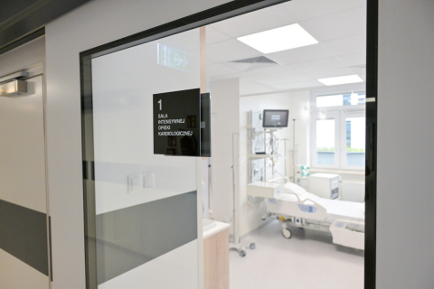 Entrance to a bright cardiac intensive care room. A black sign on the door reads “1 CARDIOLOGY INTENSIVE CARE ROOM.” Inside, white walls, a hospital bed, monitors and medical equipment. Large windows let in light.