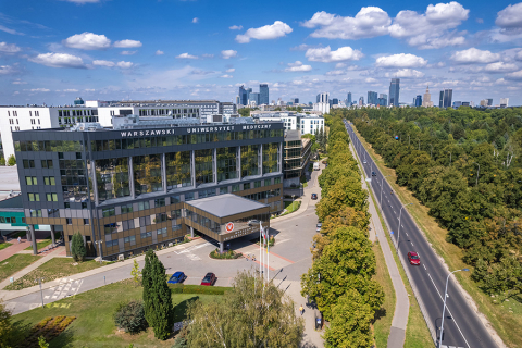 In the foreground, there is a modern building with large windows and the words “Warsaw Medical University” on the roof. There are flags in front of the building. On the right, there is a wide street surrounded by trees. In the background, there is a panorama of the city with skyscrapers and office buildings.