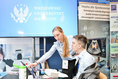 Dwie osoby siedzą przy biurku w biurze. Kobieta z długimi blond włosami wskazuje na ekran komputera. Mężczyzna w okularach patrzy na ekran. Oboje noszą niebieskie koszule i identyfikatory. Na biurku leżą chusteczki, długopisy i butelka z płynem dezynfekującym. W tle widać duży monitor i plakat z obrazem medycznym.