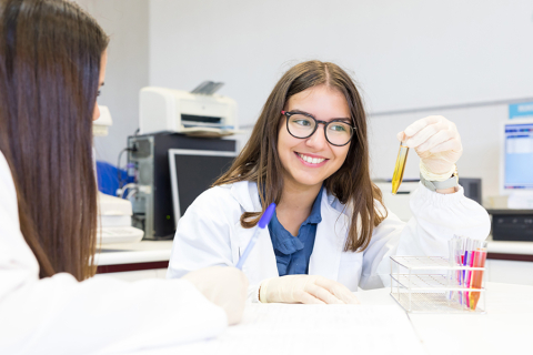 Dwie młode kobiety, ubrane w białe fartuchy, siedzą przy stole w laboratorium. Jedna z nich - zwrócona frontem do zdjęcia, trzyma w ręce fiolkę z preparatem, druga - zwrócona tyłem, zapisuje coś w notesie. Za nimi widać sprzęt laboratoryjny.
