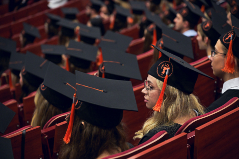Rows of people sitting in the auditorium, wearing black caps with orange tassels and black academic gowns. The caps feature round emblems with inscriptions and graphics. 