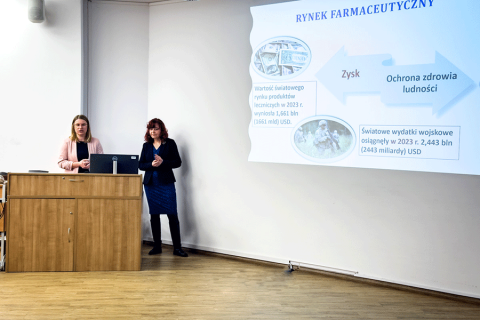 Dwie kobiety stoją w mównicy w dużej sali. Jedna osoba stoi przy komputerze, druga wskazuje na ekran z prezentacją. Na ekranie widoczny jest tytuł „Rynek farmaceutyczny” oraz schemat z napisami „Zysk” i „Ochrona zdrowia ludności”. W tle inne osoby.