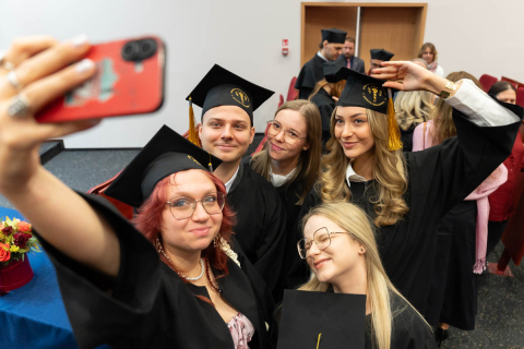 Six people wearing graduation gowns and caps. They are standing close together in a room with red chairs and wooden doors in the background. One person is holding a phone and taking a selfie. The group is posing for the photo, with some raising their hands in a gesture of posing.