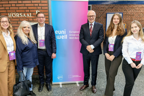 Six people standing in a room by a brick wall. In the background, there is a blue and pink banner with the inscription “euni well – European University Well-Being.