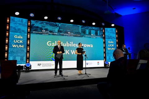 On stage during the UCK WUM Jubilee Gala, two people are standing at microphones, dressed in elegant attire. In the background, there is a large screen with a blue graphic displaying the following inscriptions: “UCK WUM Jubilee Gala,” “125th Anniversary of the Infant Jesus Clinical Hospital UCK WUM,” “50th Anniversary of the Central Clinical Hospital UCK WUM,” and “10th Anniversary of the Opening of the New UCK WUM Building.”