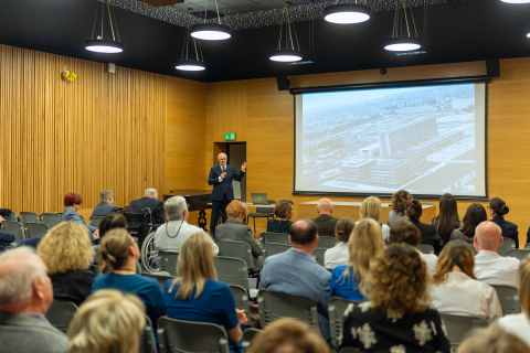 A large hall where several dozen people are gathered. Everyone is seated except for Professor Rafał Krenke, who is standing in the middle of the room and speaking into a microphone. Behind him, a large multimedia screen is visible.