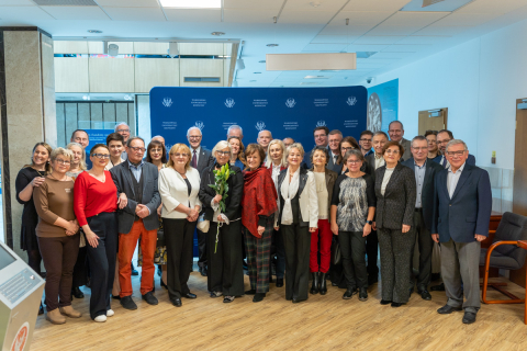 Grupa emerytowanych pracowników oraz władze uczelni pozują do zdjęcia grupowego. Wszyscy się uśmiechają. Za nimi widać niebieską ściankę z logotypwem WUM.