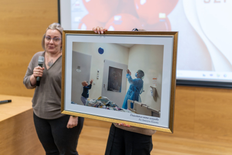Two people are standing next to each other; one is holding a microphone, the other a large framed photograph showing a child playing with a balloon in a hospital room with an adult in protective clothing. 