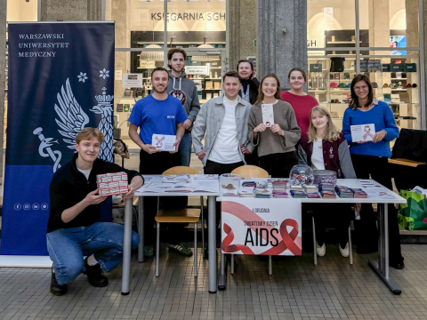 Grupa studentów stojących przy stoisku informacyjnym z ulotkami, materiałami edukacyjnymi i gadżetami promującymi zdrowie w przestrzeni uczelni.