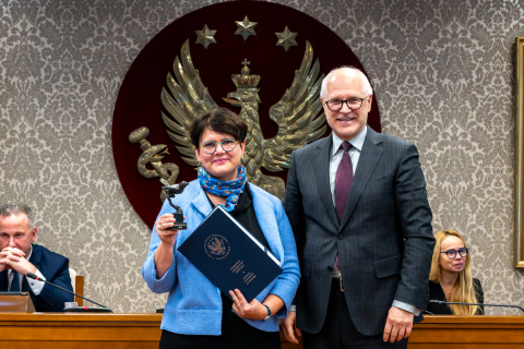 On the left, Prof. Joanna Kolmas, wearing a blue jacket, holds a cuckoo statuette in her raised right hand and a navy blue briefcase in her left. On the right is the rector. Both are smiling for the camera. Two other people can be seen in the background.