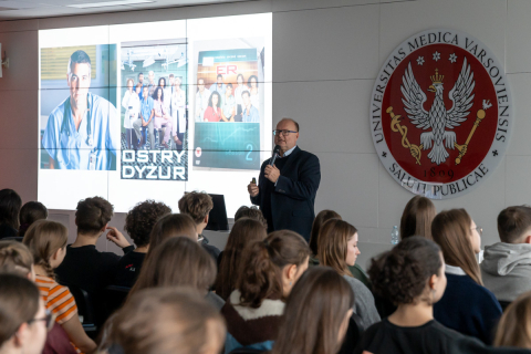 Prof. Czupryniak w czasie wykładu stoi na środku sali. Za nim widać czerwone godło WUM oraz ekran multimedialny z wyświetlonymi zdjęciami. Przed nim w rzędach siedzą młodzi ludzie.