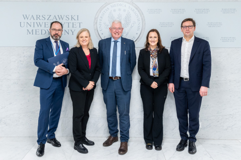 Pięć osób, elegancko ubranych: trzech mężczyzn i dwie kobiety. Wszyscy się uśmiechają, pozując do zdjęcia.