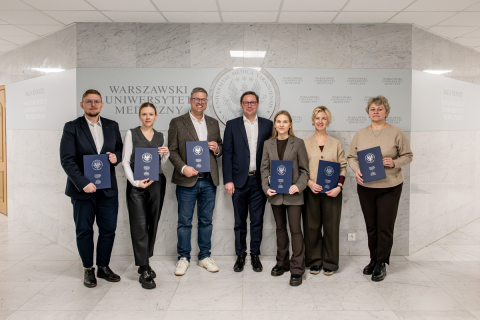 Seven people are posing for a photo. They are dressed formally. Six of them are holding navy blue folders with diplomas. They are standing in a bright room.
